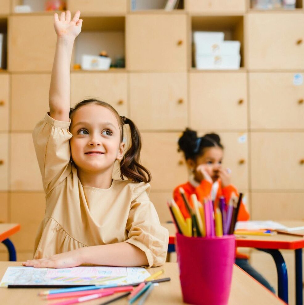 Three children engaged in learning activities inside a classroom, with one raising her hand.